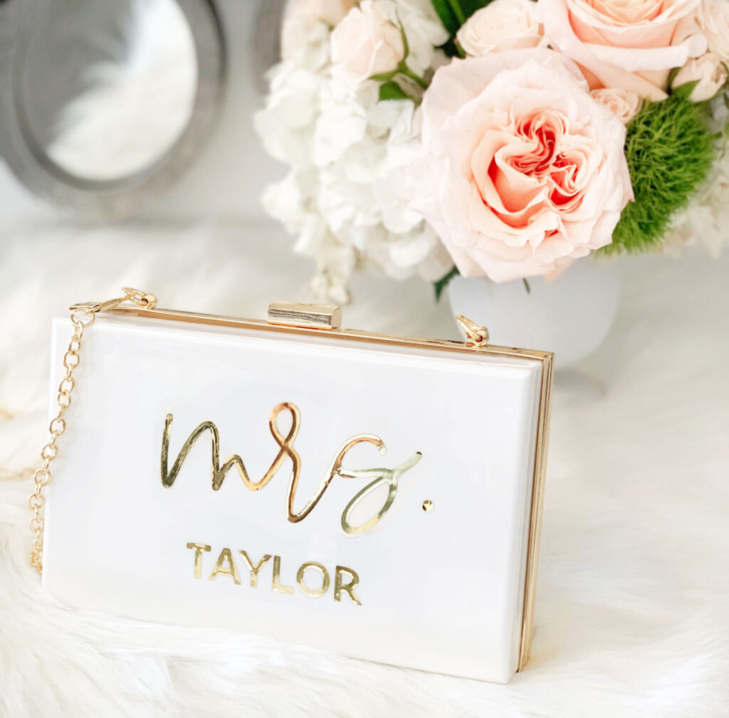 In the the middle stands a beautiful white and gold acrylic clutch purse with large text "mrs." and smaller text featuring the brides last name underneath makes this a stunning option and best gift for the bride.