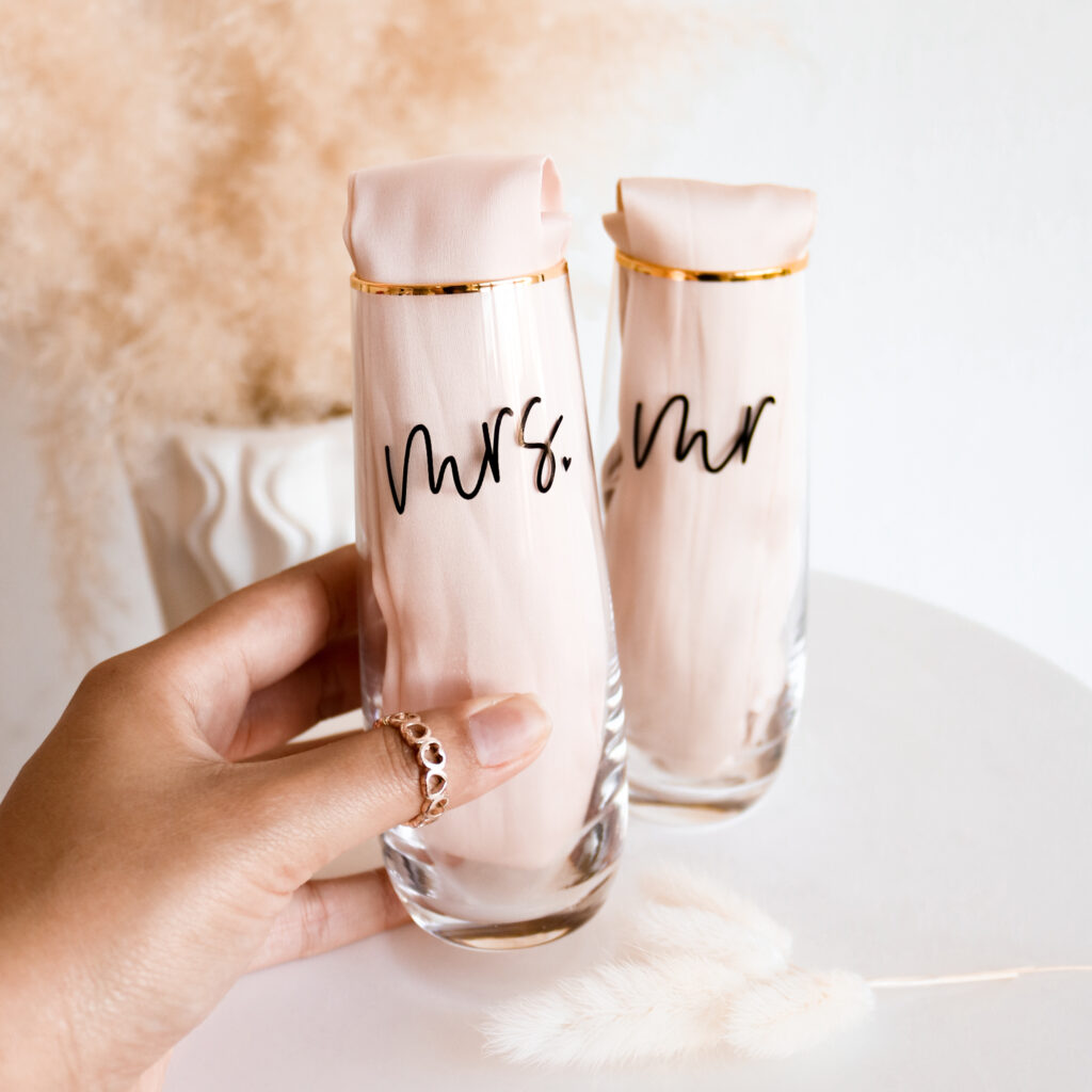 A light colored hand is holding a glass champagne flute with a gold rim that has black text saying "mrs." Behind the "mrs." glass is a matching "mr." glass. 