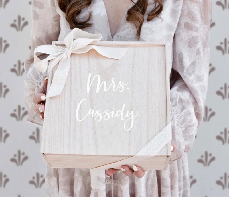 A woman is holding a light colored square wooden box with a baby pink ribbon wrapped around the corners and has a personalization in the middle written in white that says "Mrs. Cassidy" makes a wonderful gift for the bride!