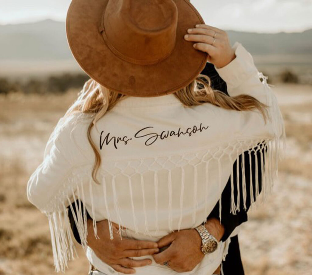 Bride wearing white fringe jacket that showcases her new last name standing in the desert with her husband. Her outfit is for reader inspiration for creating a disco cowgirl outfit.