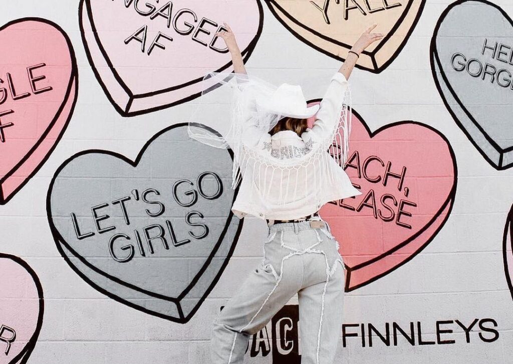 Bride dressed in white fringe jacket, white cowboy hat, and blue jeans, is raising her arms up in excitement while standing in front of a wall painted with a colorful candy hearts mural.